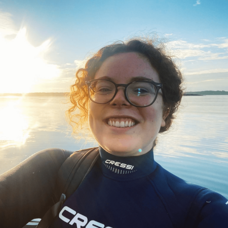 Bridget is standing in the sea wearing a wetsuit. They have curly hair and are wearing glasses. The sun is glowing in the background.